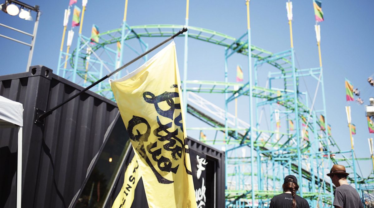 Yellow Buck the Quo flag hanging from black shipping container in front of rollercoaster